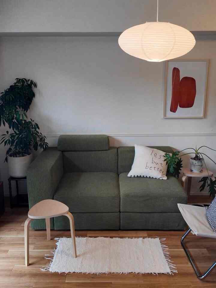 ナチュラルな色調のリビングルーム、緑のコーデュロイソファと木製のスツール、植物、丸い和紙のペンダントライトが配置された雰囲気のある空間