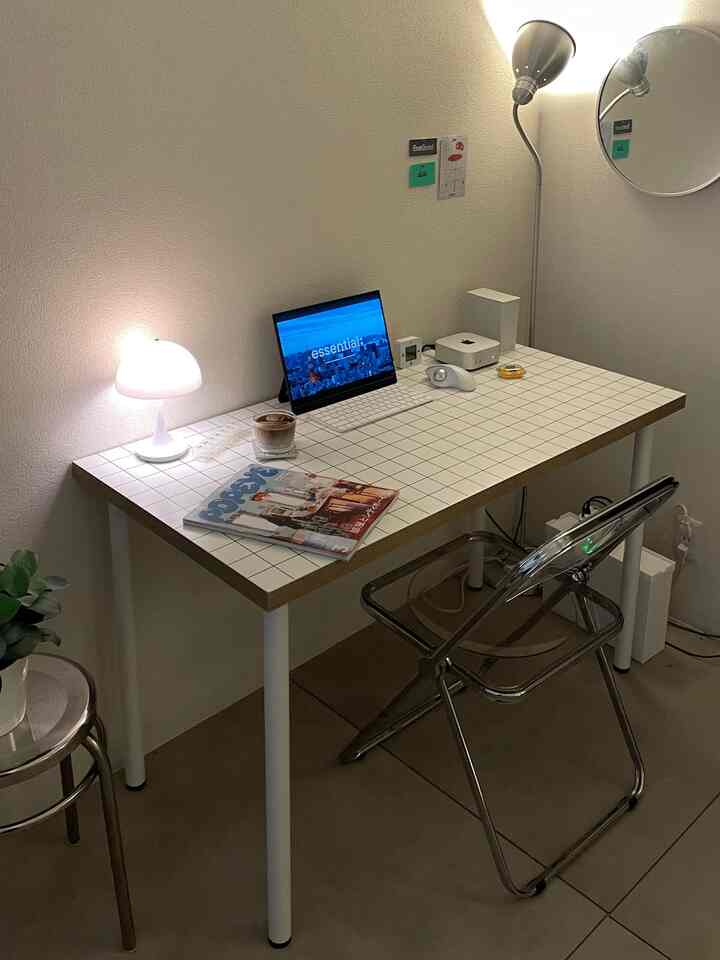 白とシルバーのトーンのワンルームホームオフィス、タイル模様のデスクと透明なスツール、照明が配置された清潔感のある空間