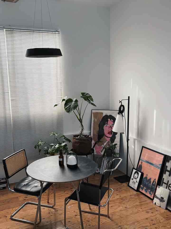 モノトーンのミッドセンチュリーモダンなダイニング、丸いテーブルとラタンチェア、複数のポスターと観葉植物が配置されたシンプルな空間