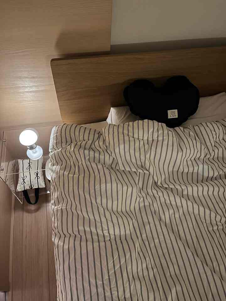 温かみのあるベージュと木目調の1LDK寝室、ストライプ柄の寝具、ナイトテーブル、クッション、間接照明が配された雰囲気のあるインテリア