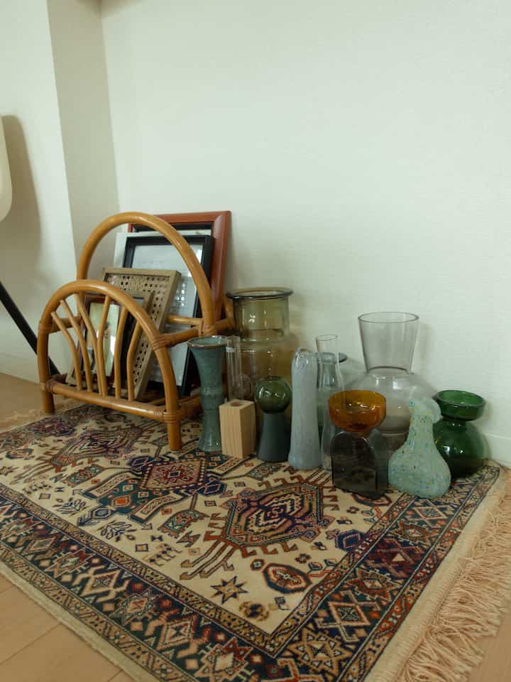 ベージュとブラウンのトーンが調和するリビングの一角、籐のマガジンラックと色とりどりのガラス製花瓶が配された雰囲気のある空間