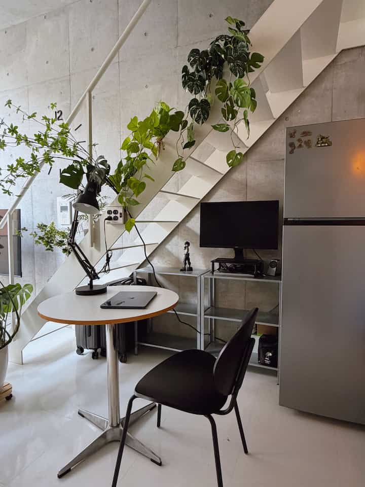 白い床と階段下のシルバー棚、緑の植物が配置されたコンパクトでモダンなホームオフィスのワークスペース
