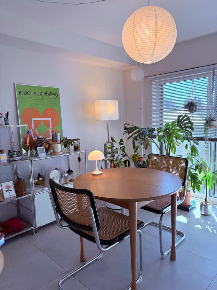 白い壁と木目調の丸テーブル、ラタンチェアが配置されたミッドセンチュリーモダンスタイルのダイニングルーム、自然光と植物が調和した印象的な空間