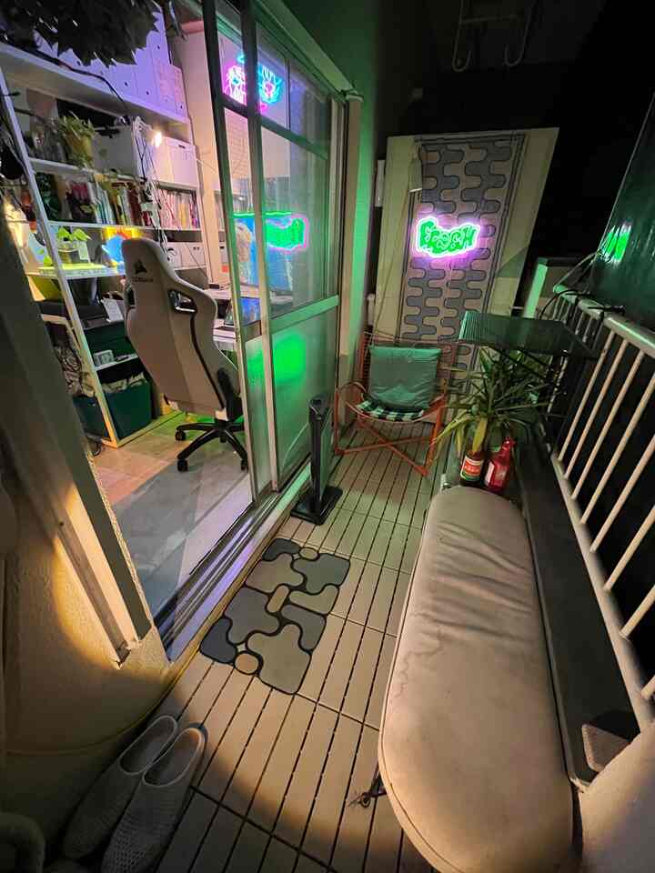 Dimly lit studio apartment balcony with white and green tones, featuring a gaming chair and outdoor chair creating a cozy atmosphere