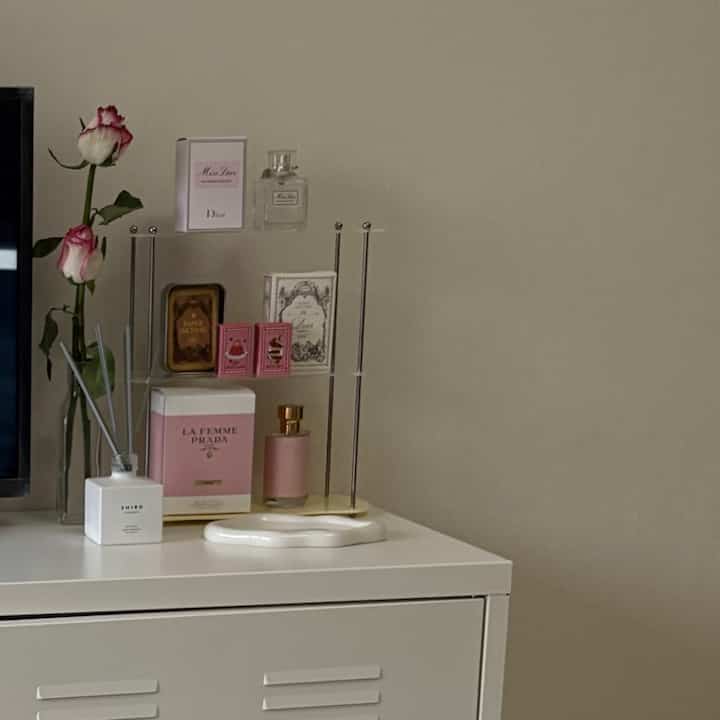 Beige wall with a white cabinet featuring Prada and Dior perfumes and rose vase objets in a clean, minimalist space