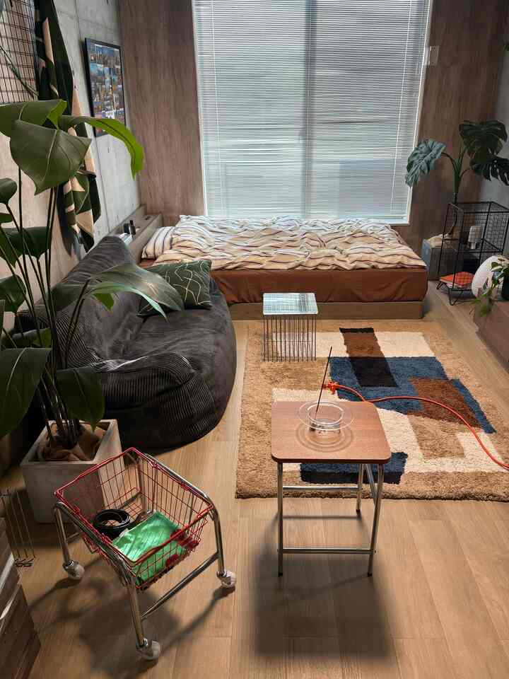 ウッドトーンの壁と床の1Kリビング兼寝室、ダークグレーのソファとスツール、植物が配置された雰囲気のある空間