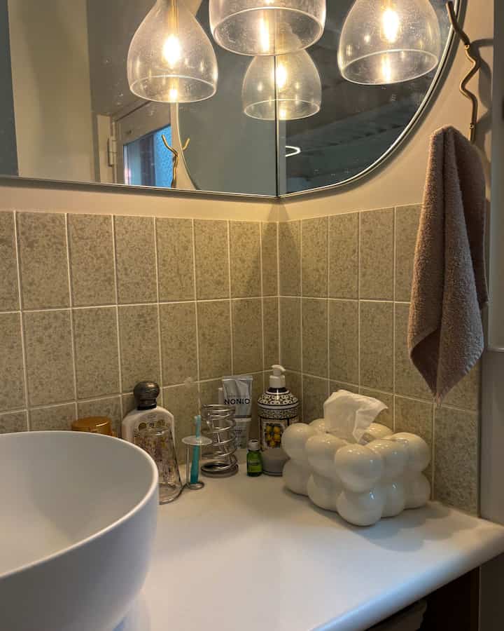 ベージュのタイル壁と白いセラミックシンク、ゴールドの照明とタオルが配置されたバスルームの洗面台スペース