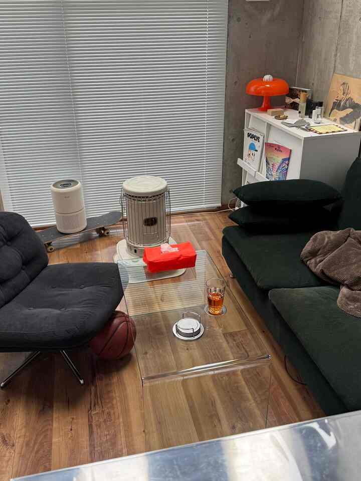 ナチュラルな木目調の床とグレーのコンクリート壁、ブラインドがあるモダンな一人暮らしリビング、黒のアームチェアと緑のソファを配置