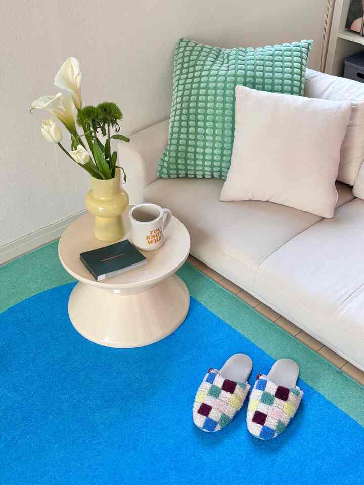 パステル調の1LDKリビング、白いソファとクリーム色の丸いスツールテーブル、青いラグと花瓶が配置されたナチュラルな空間