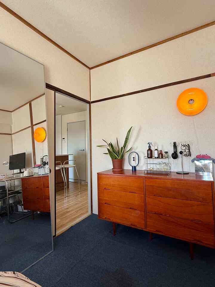 ベージュの壁と木製のドレッサーが置かれたコンパクトな玄関空間、オレンジのブラケットライトと植物が調和したナチュラルなインテリア