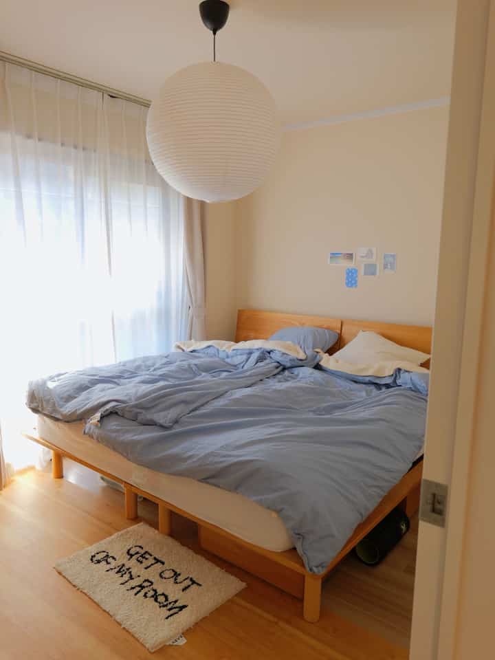 ベージュと木目調が調和する静かな寝室、窓際に青い寝具と小さなラグが配置された落ち着いたインテリア