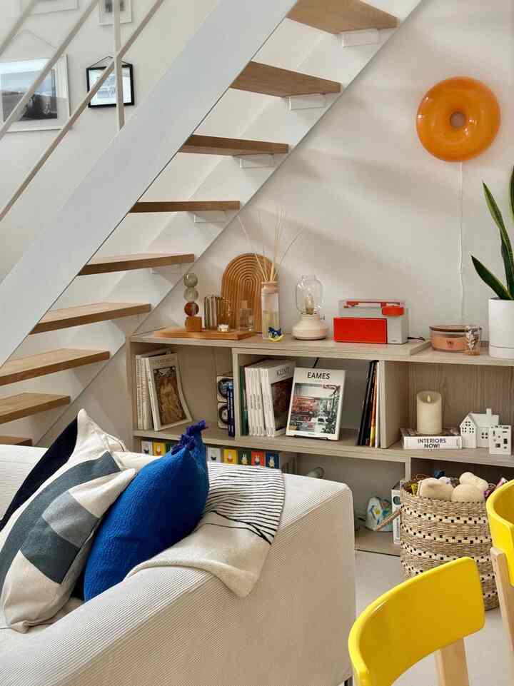 Natural tone living room featuring a white sofa with blue cushions, wood-tone stairs, and a bookshelf creating cozy ambiance