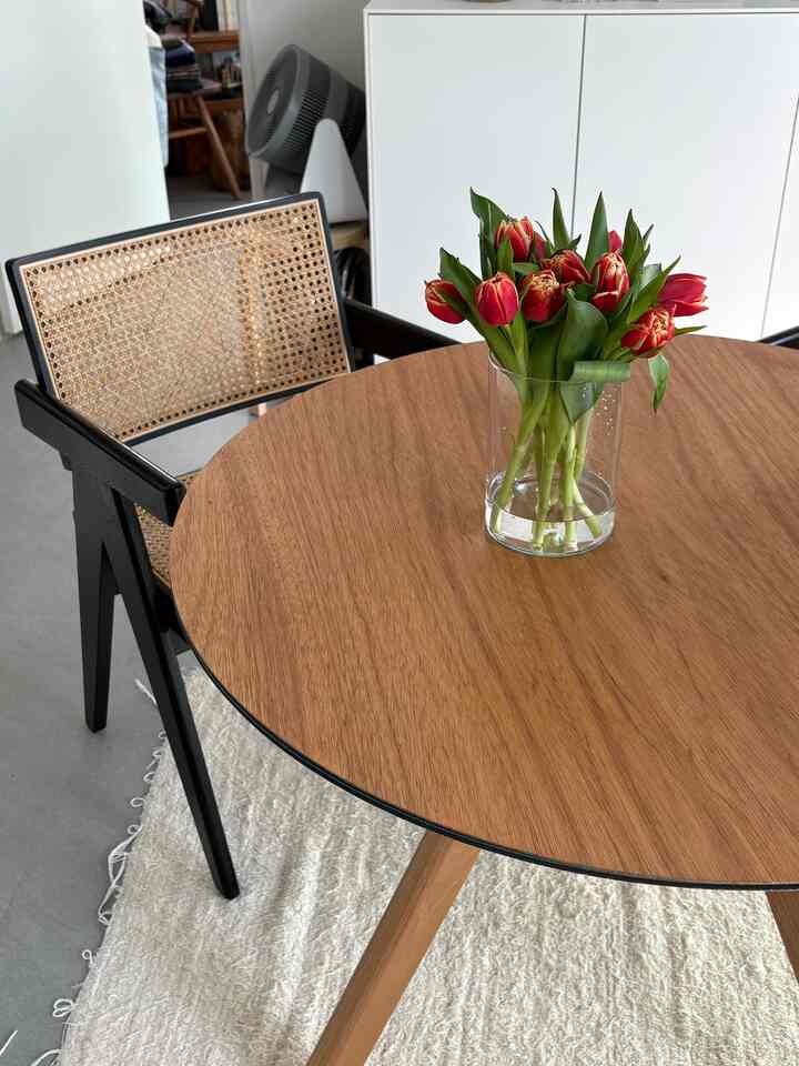 ナチュラルトーンの木製丸テーブルと黒いラタンチェアが配置され、透明な花瓶に赤いチューリップが飾られた雰囲気のあるダイニング空間