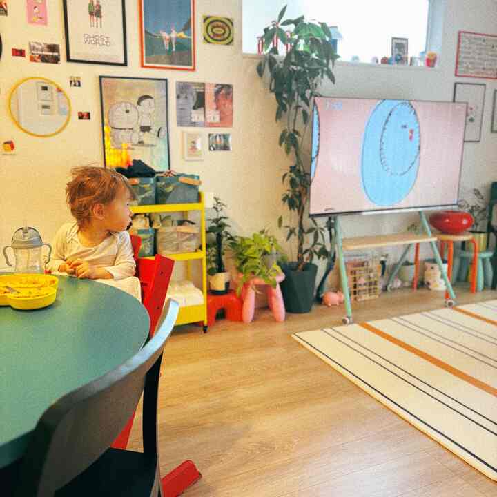 明るいナチュラルトーンのダイニングルーム、子供がテーブルに座り、テレビと壁のアートが配置された温かみのある空間