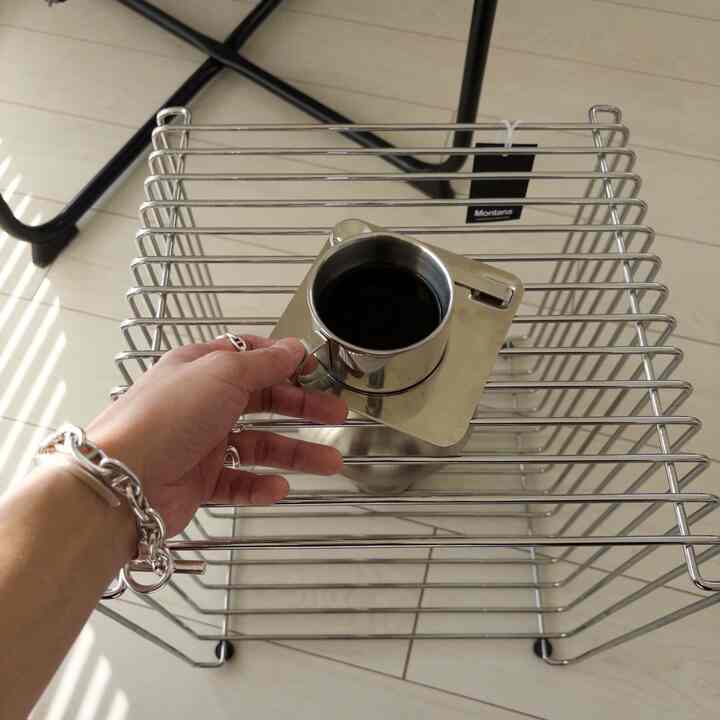 シルバーの金属ワイヤーシェルフの上にコーヒーカップを持つ手が見えるシンプルな空間