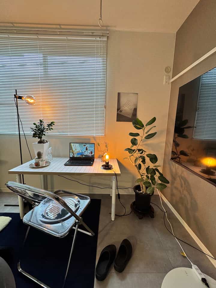 白とベージュの色調のワンルームホームオフィス、透明な椅子と植物、デスクランプが配置されたモダンな作業空間