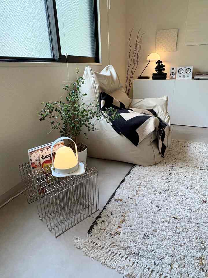 ナチュラルな色調の小さなリビング、ベージュの大きなビーズクッション、白いラグ、棚とフロアランプが配置された雰囲気のある空間