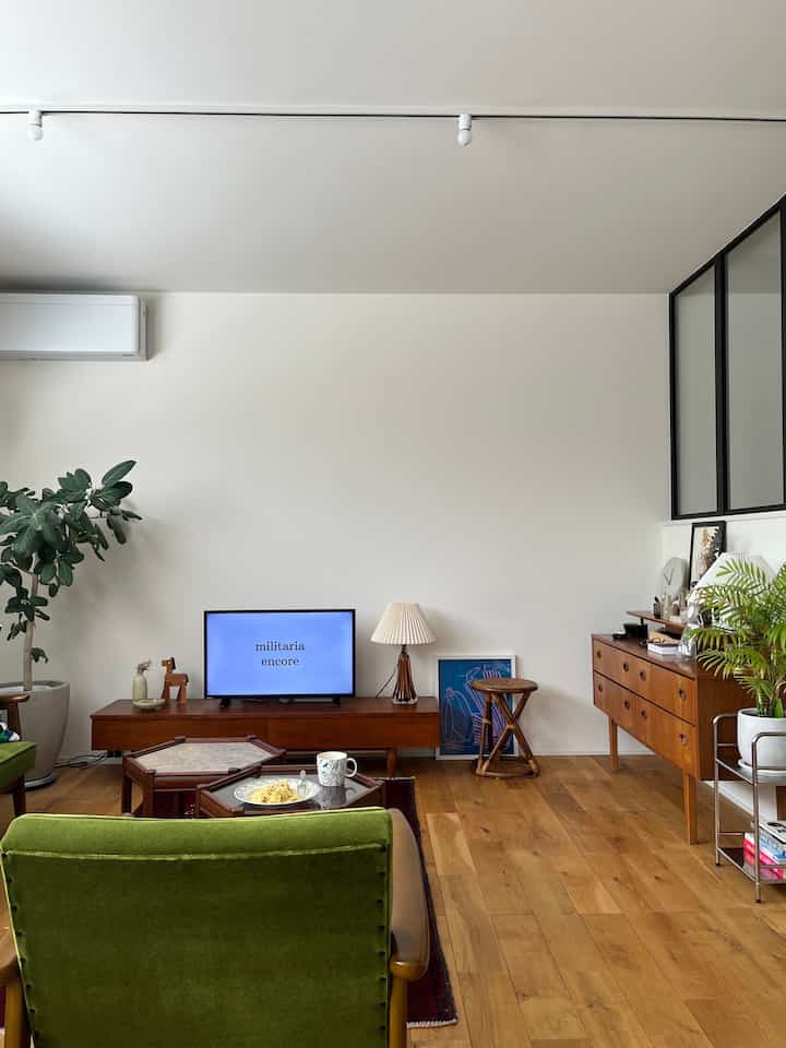 A living room with white walls and wood-toned floor, featuring vintage mid-century modern furniture and large indoor plants creating a cozy atmosphere