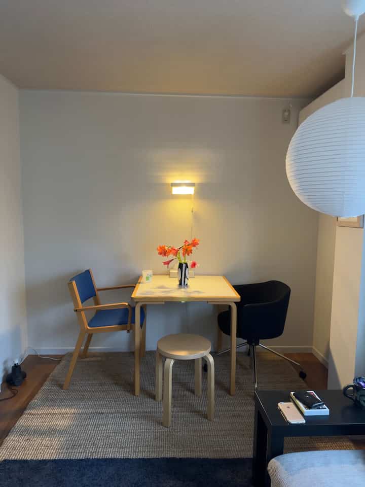 Natural oak-toned dining table and chairs in a simple two-person dining room, featuring wall light and flower vase creating cozy atmosphere