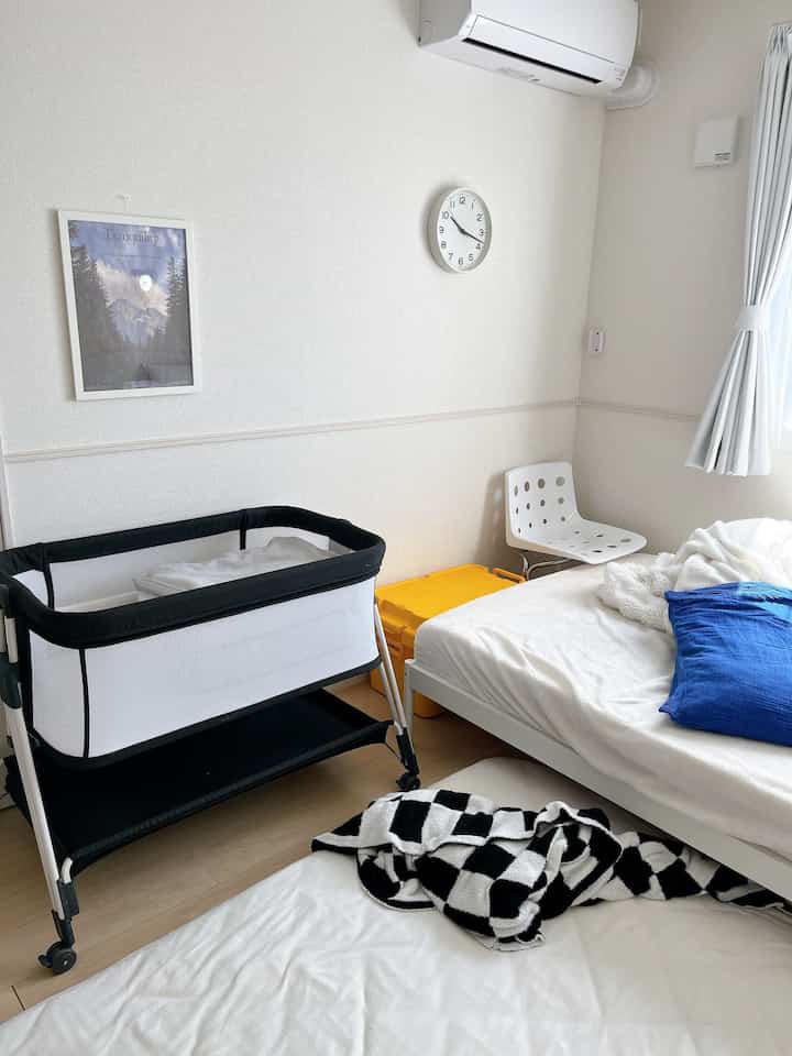 白とナチュラルな色調の寝室、ベビーベッドとベッド、壁掛け時計が配置された落ち着いた朝の空間