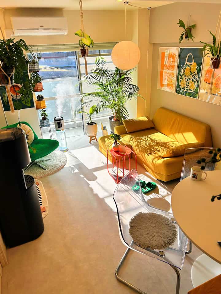 黄色いソファが中心のリビング、緑の植物と丸いペンダントライトが配置された温かみのある明るいインテリア