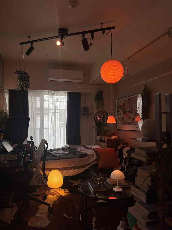 Warm orange-lit 1K bedroom and home office space featuring desk, bed, and plants in a cozy interior