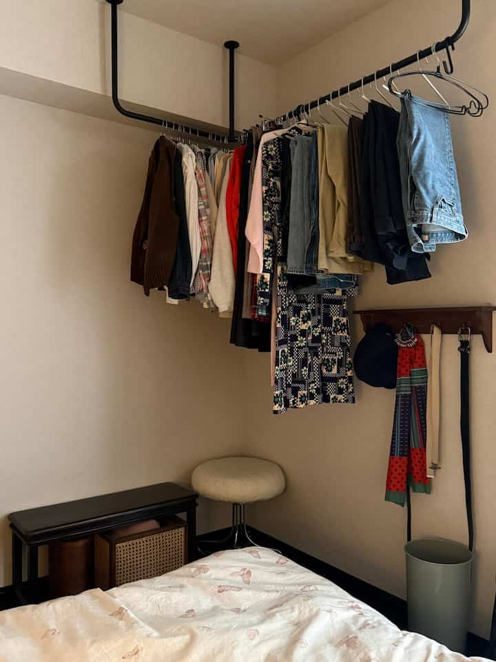 ベージュの落ち着いた色調の小さい寝室、クローゼットに掛かった服と居心地の良いベッドカバー、スツールを備えた雰囲気のあるインテリア