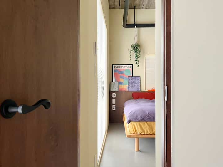 Beige walls and wood tone furniture define a small single bedroom featuring a wooden bed frame, purple bedding, wall sconce, and hanging plant for a cozy atmosphere