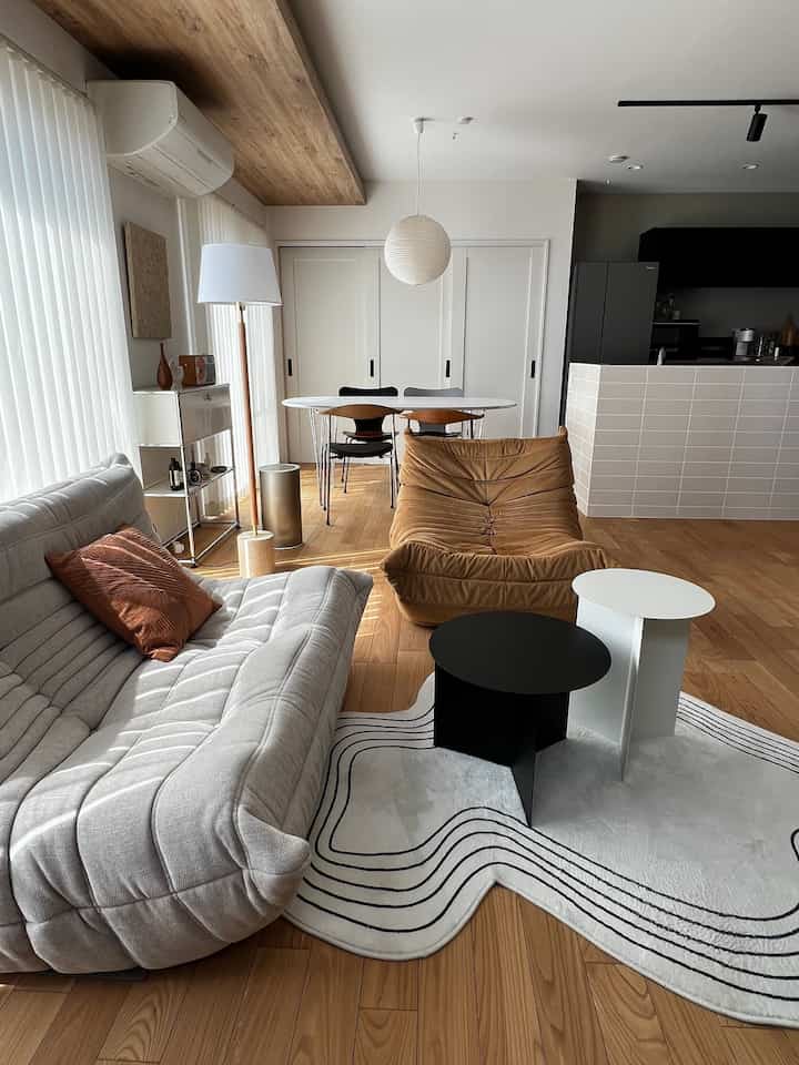 A spacious and bright mid-century modern living room featuring natural wood tones, white walls, a comfortable sofa, armchair, and a modern dining table.