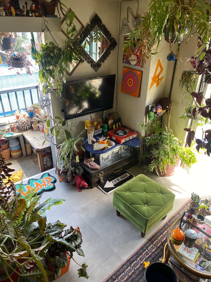 白い壁と多様な緑の植物が配置されたレトロ風リビング、オットマンとレコードプレーヤーがある印象的な空間