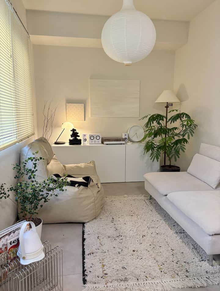 White-toned living room featuring sofa, beige bean bag, floor lamp, and plants in a simple natural space