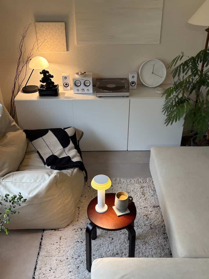 Simple modern bright living room featuring cream sofa, dark brown stool, white storage cabinets, table lamp, and plants creating cozy ambiance