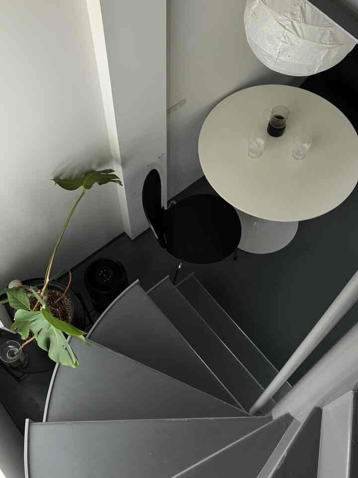 ホワイトとダークグレーのトーンのコンパクトな空間、丸テーブルと黒い椅子、階段と観葉植物が配置されたモダンなインテリア