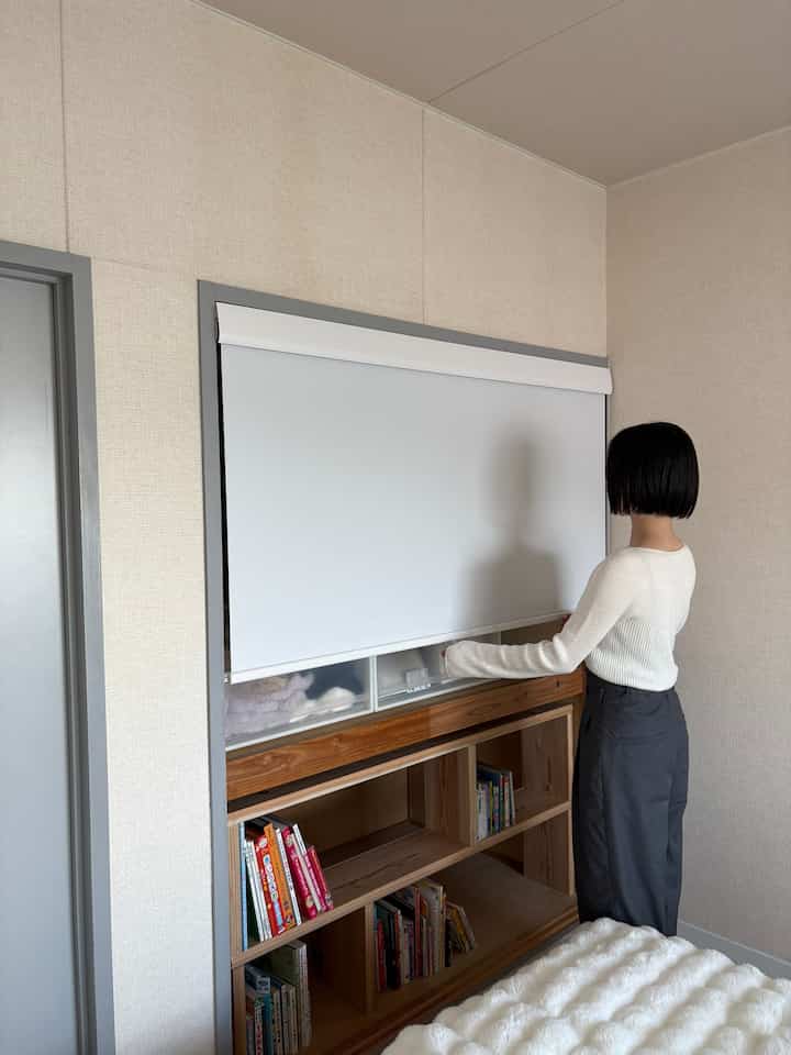 ベージュの壁と白いロールスクリーンが設置された狭い部屋、ロータイプの木製収納が配置されたナチュラルな雰囲気のインテリア