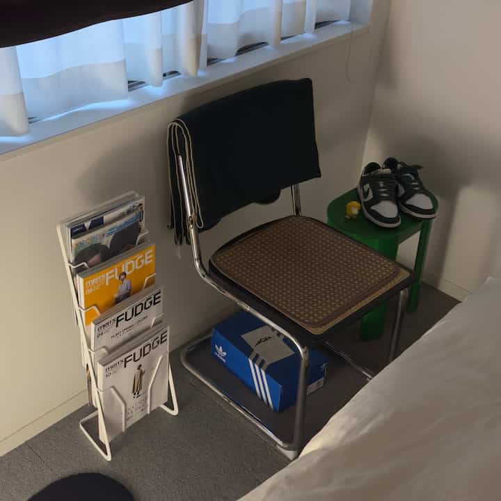 Narrow bedroom corner with white curtains, green stool, rattan seat chair, and a magazine rack beside it
