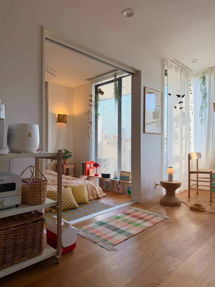 A compact bedroom and workspace with white walls and natural wood flooring illuminated by natural light, creating a cozy atmosphere