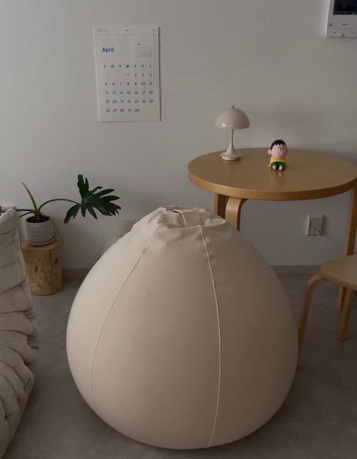 Beige-toned living room featuring a natural wood round table, a large bean bag, and a plant in a simple interior