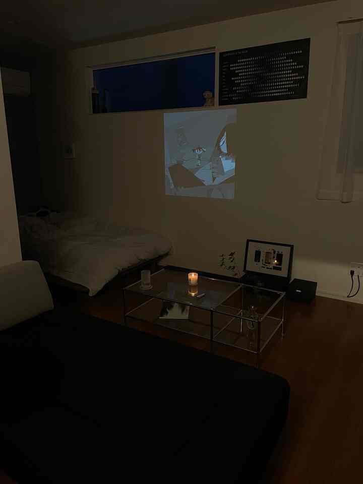 Modern living and bedroom space featuring a white bed, black sofa, glass coffee table, and a lit candle creating cozy ambiance