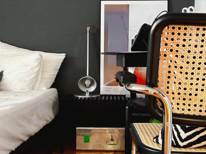 Modern bedroom with black accent wall and white bedding, featuring a rattan armchair and a metallic table lamp creating a sleek atmosphere