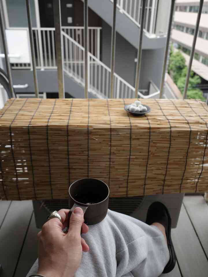 Gray and wood-tone balcony space featuring a bamboo blind and a person holding a coffee cup, creating a relaxed atmosphere