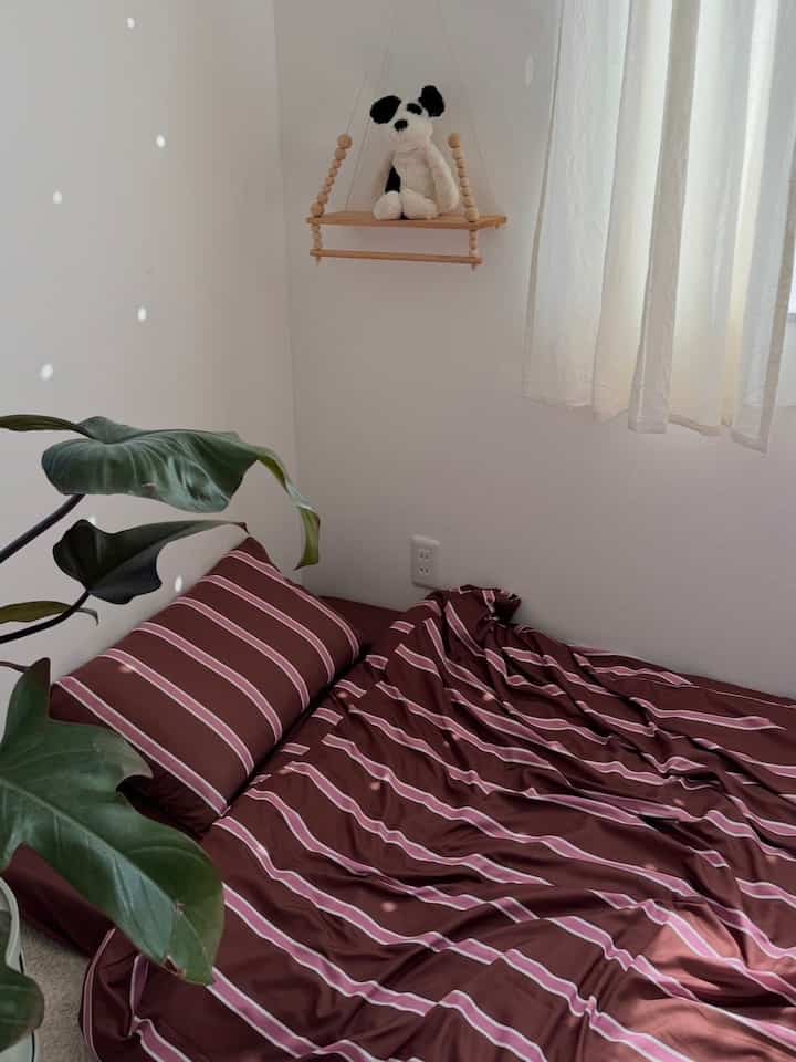 A cozy single household bedroom with white walls and brown striped bedding, featuring wooden shelf and soft natural light