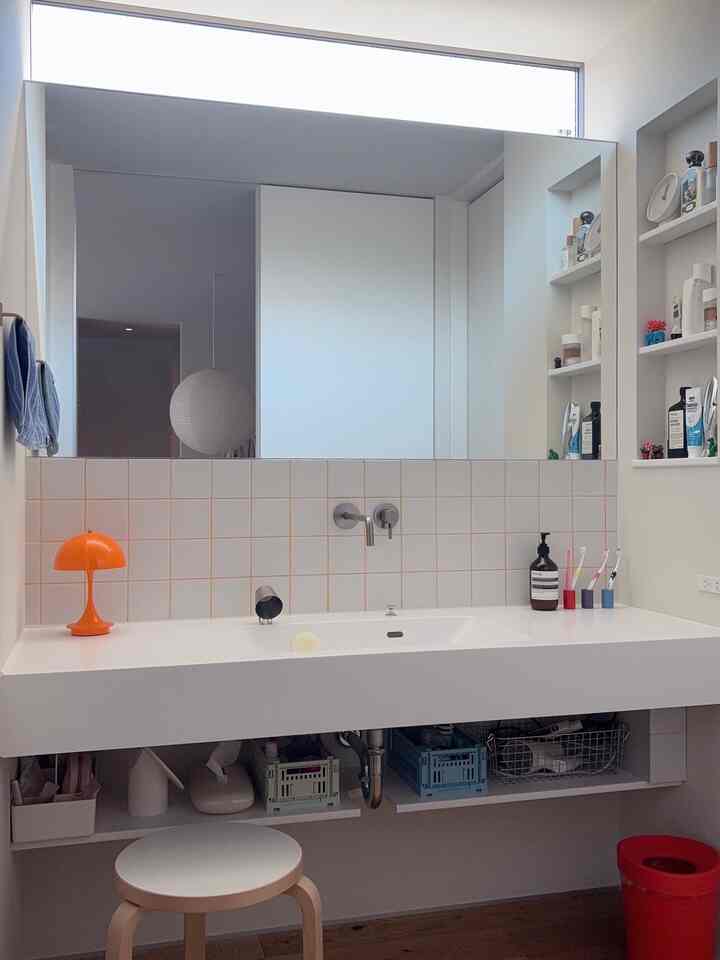 White and beige toned powder room featuring a large mirror, ceramic sink, wooden stool, and an orange table lamp harmoniously arranged