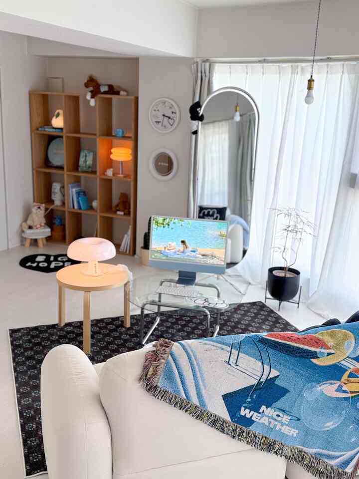 A natural-toned living room with white walls, wood furniture, sofa, bookshelf, and a computer monitor, presenting a clean, simple ambiance