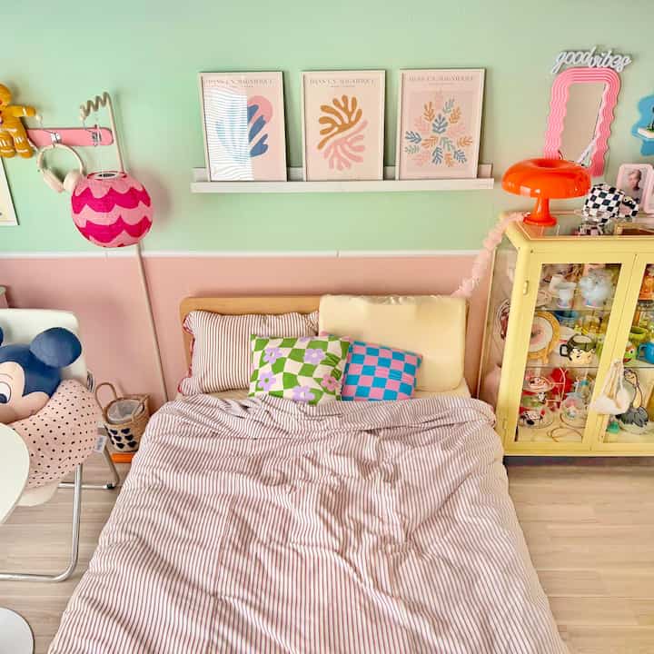 ミント色の壁とピンクの下半分の壁がある寝室、カラフルなクッションとおもちゃが置かれたベッド、黄色い収納キャビネットが配置されたかわいらしいインテリア