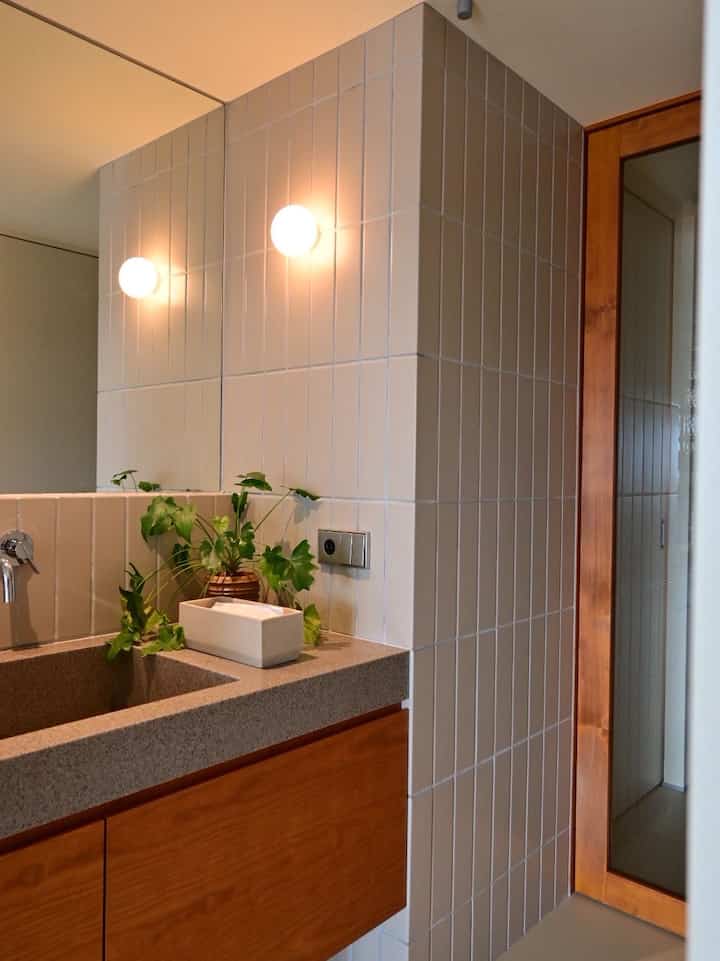 Natural style bathroom with gray tiled walls and wooden cabinets, featuring plants on the wash basin and wall sconces creating a cozy atmosphere