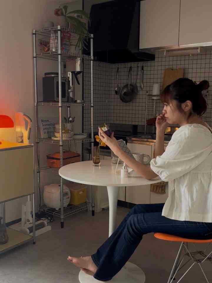 White and orange toned small 1DK kitchen featuring round table, metal shelving, and a seated woman