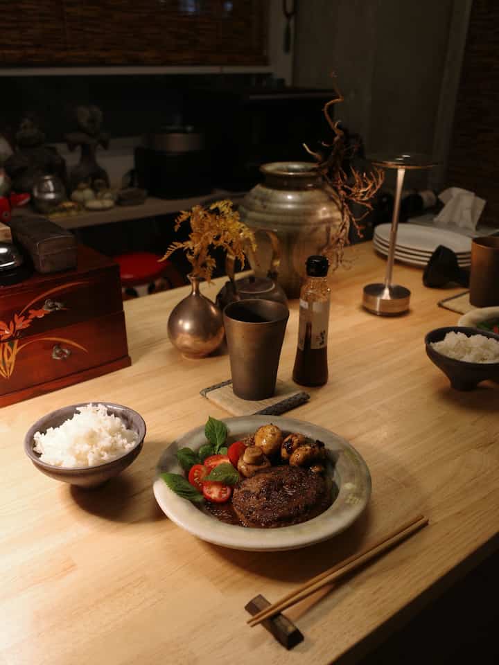 Natural-toned 1LDK dining room featuring a wooden dining table with homemade meal, traditional ceramics, and a table lamp creating cozy atmosphere