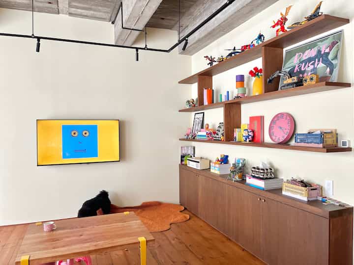 Natural wood-tone living room featuring wall-mounted TV, shelves with toys, and a visible black dog creating a cozy atmosphere