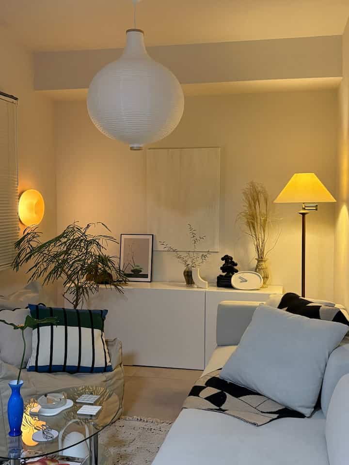 Warm-lit simple modern living room in white tones, featuring green plants and cushions for a cozy atmosphere