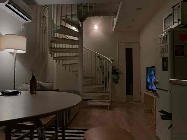 Dimly lit studio apartment dining room with white walls and a spiral staircase, featuring a wooden table and sofa creating a cozy vintage atmosphere